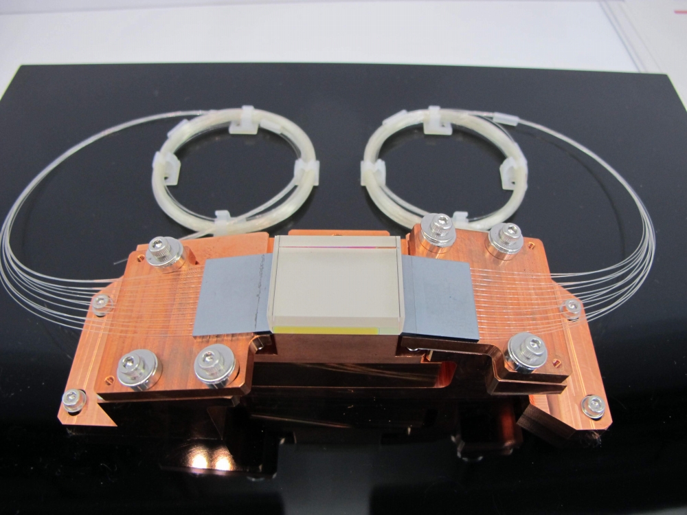 システムの心臓部「アイセーフ高出力導波路型レーザー増幅器」。銅のヒートシンクが物々しさを感じさせる、ハイパワーのレーザー発振器である。中央の四角いガラス部分に導かれたレーザー(波長1531.4nm)を、周囲のファイバー群から送り込むレーザー(波長940nm)でパワーアップさせる。増幅のカギとなるのはガラスに添加されたエルビウム(Er)という元素。深い海の底でインターネットを支えている、光海底ケーブル網の光増幅中継機と同じ原理。最近では小型で航空機搭載可能なドップラーライダーも開発され、ニュースになっている(JAXAとボーイングが共同開発中)が、そのユニットも三菱電機製という